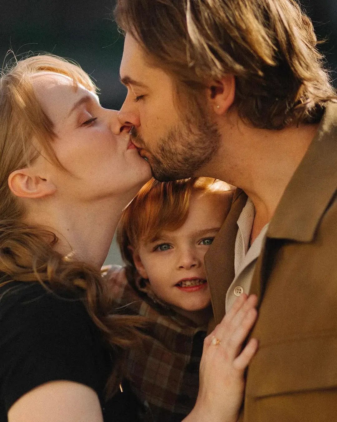 A couple kiss in warm evening light while their young red-haired child stands between them, looking at the camera.