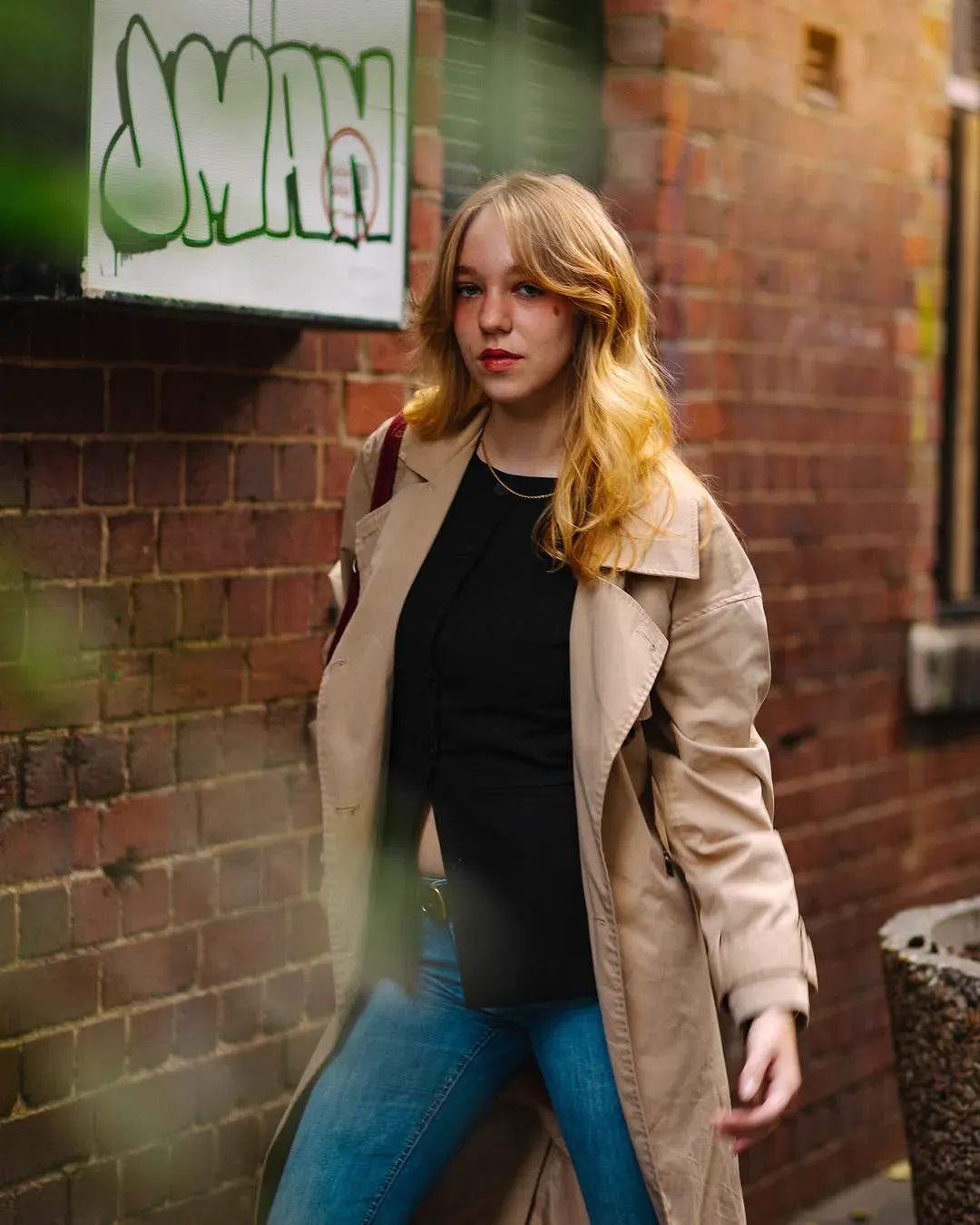 A young woman in a beige trench coat stands against a red brick wall tagged with graffiti, looking past the camera.