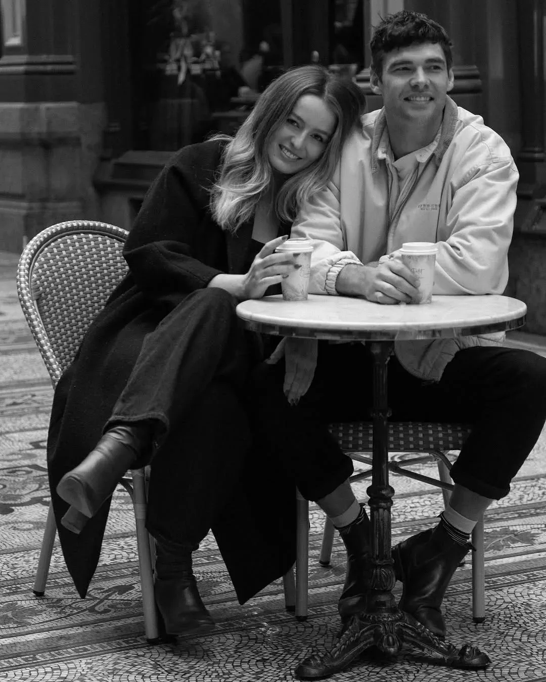 Black and white photograph of a couple seated at a small marble cafe table with takeaway cups, leaning into one another.