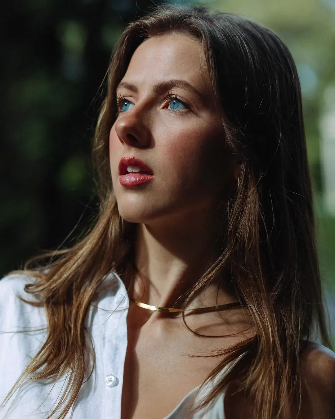 Close portrait of a woman in a white shirt looking upward, dappled sunlight across her face and a fine gold chain at her throat.