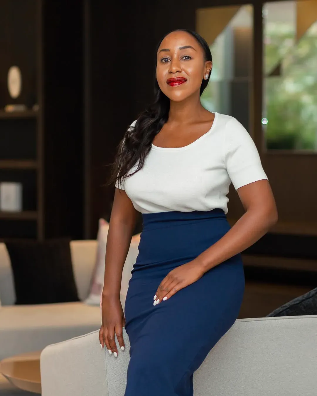 A woman in a white top and navy pencil skirt leans against a pale sofa in a dark, softly lit interior.