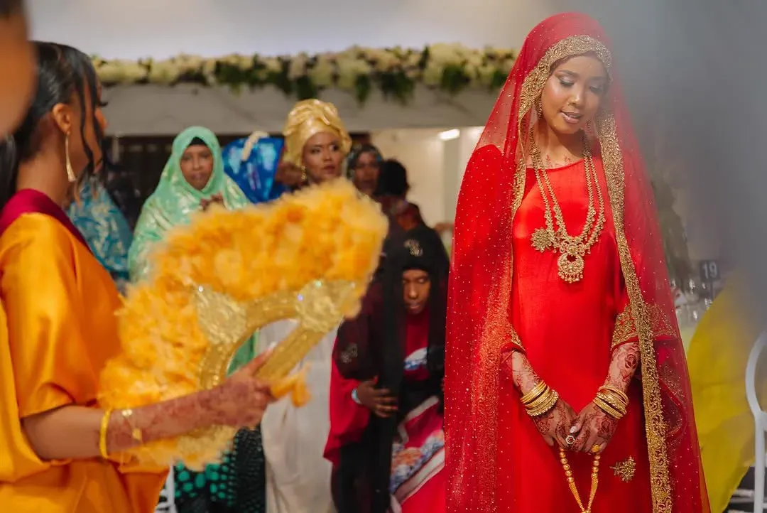 A bride in a red and gold veil and heavy gold jewellery walks through her wedding reception as guests look on.