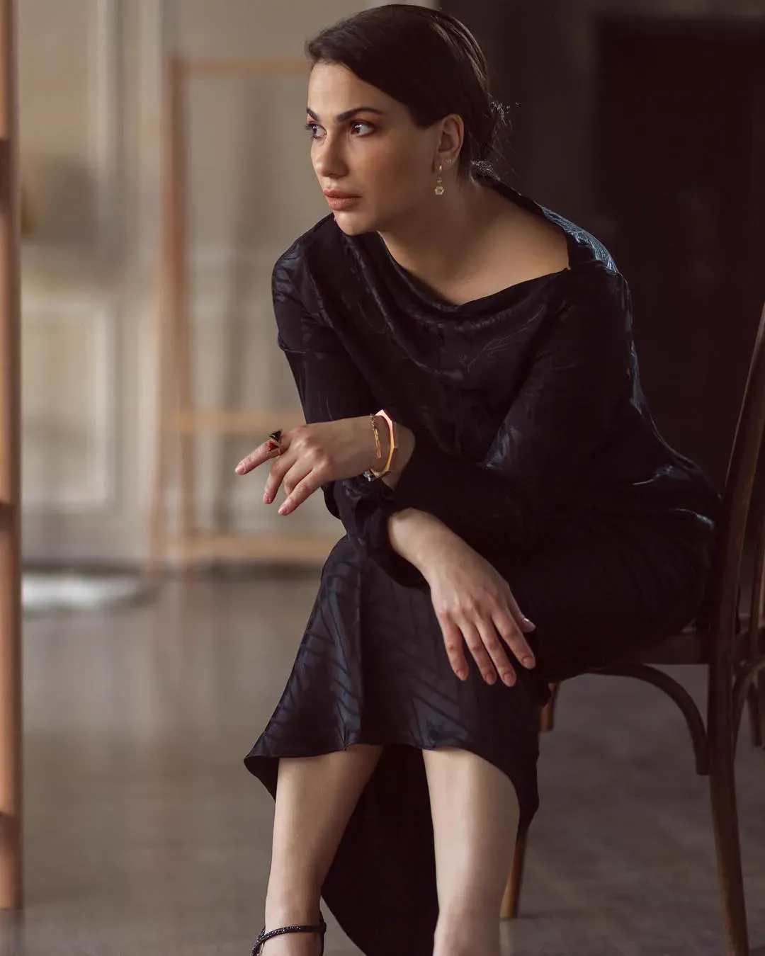 A woman in a flowing black dress sits sideways on a wooden chair in a bare, sunlit room, looking off to one side.