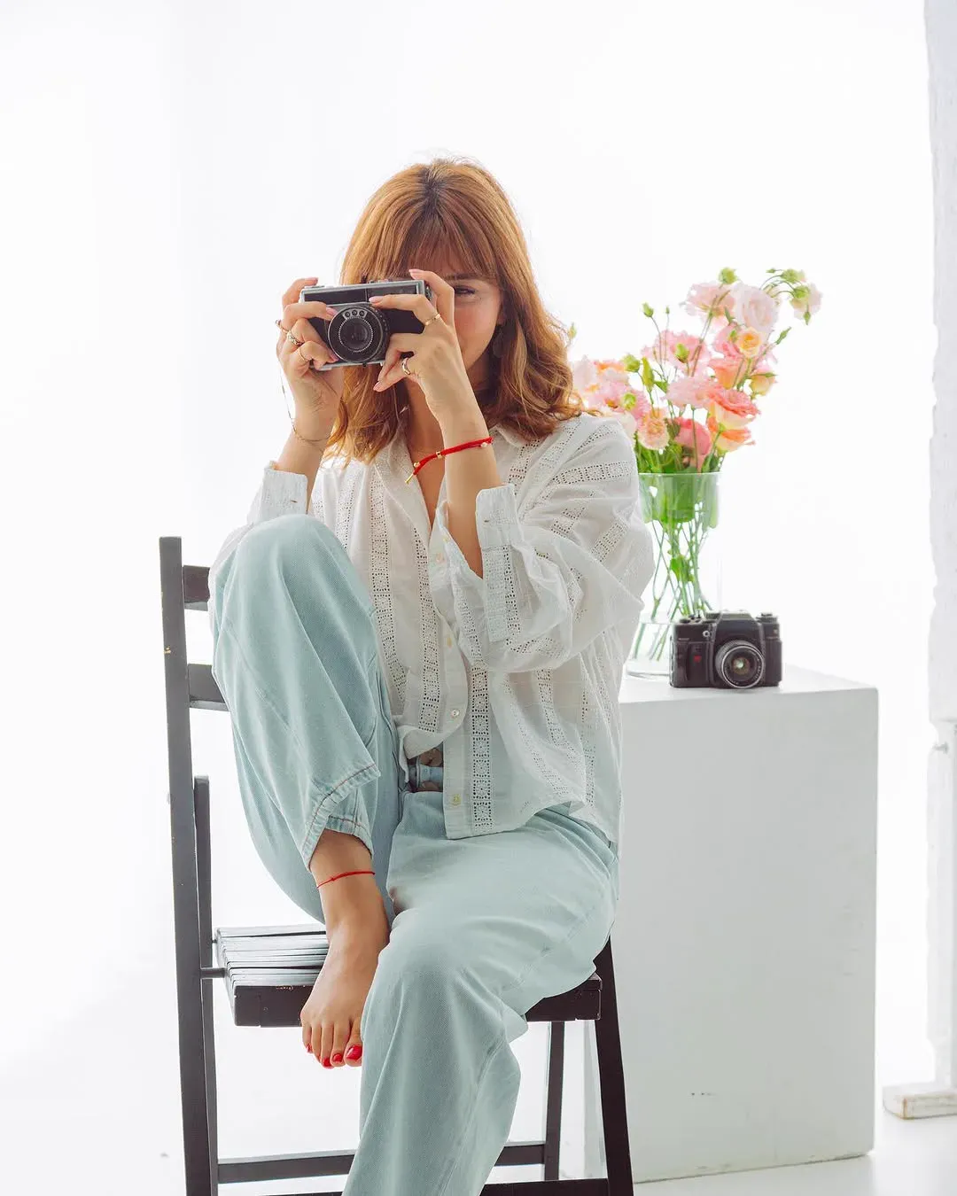 A woman sits barefoot on a chair in a white studio raising a film camera to her eye, a vase of pink roses beside her.