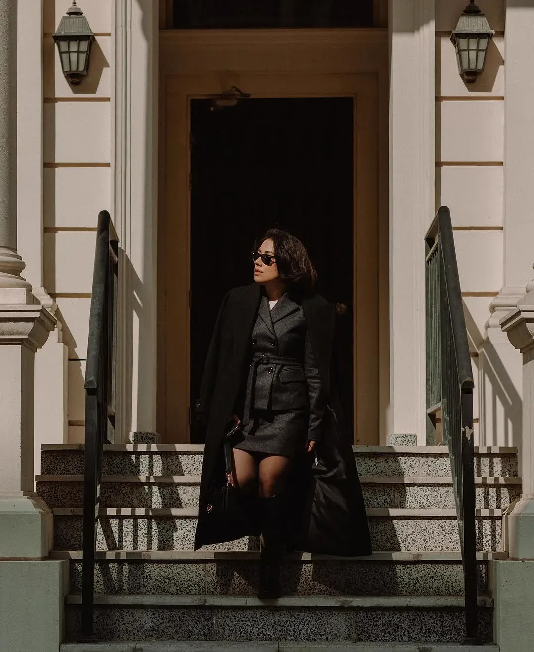 A woman in a dark coat and sunglasses descends the steps of a neoclassical building in hard afternoon light.