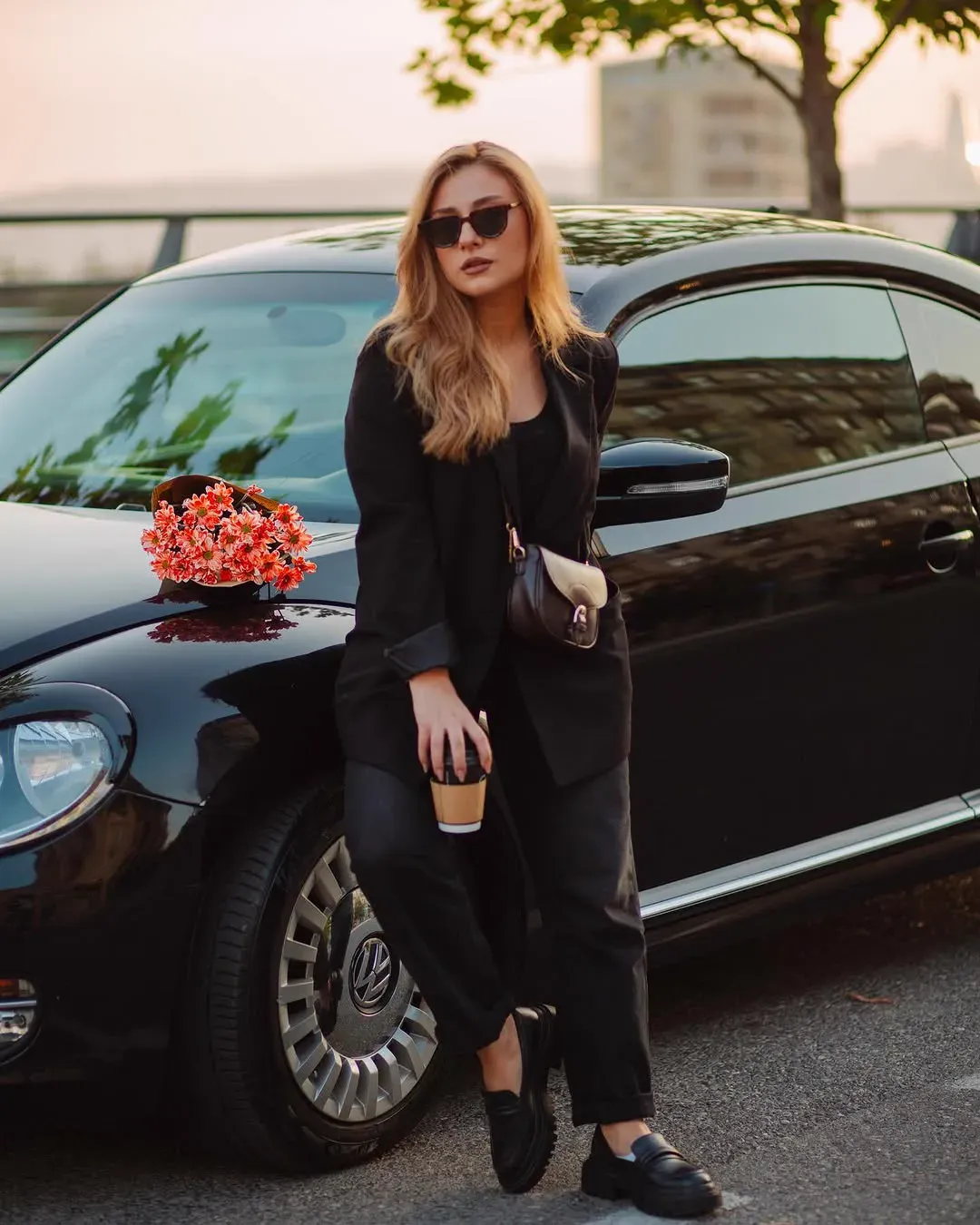 A woman in black leans against a black Volkswagen Beetle at sunset holding a coffee cup, flowers resting on the bonnet.
