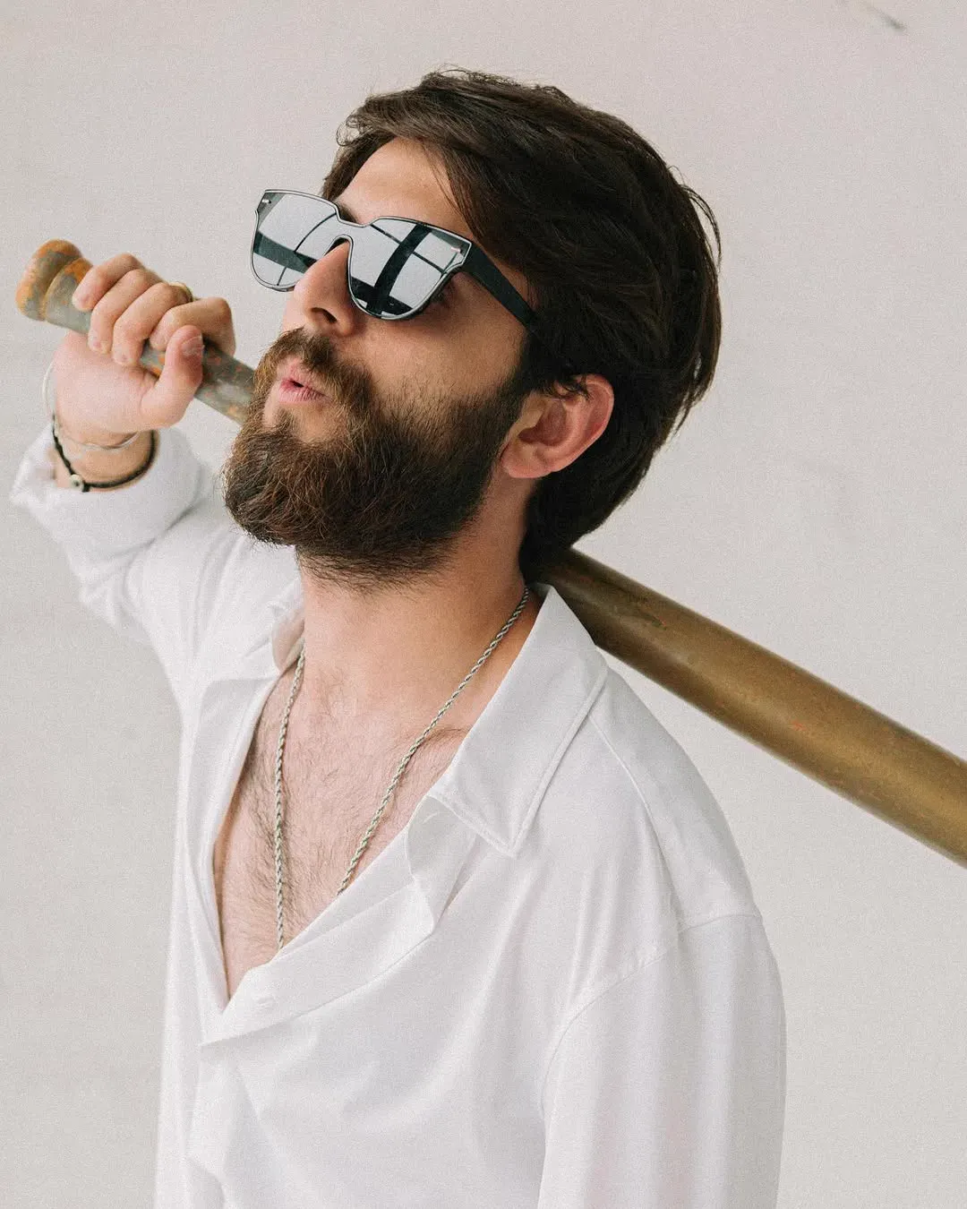 A bearded man in an open white shirt and angular sunglasses rests a wooden bat across his shoulder.