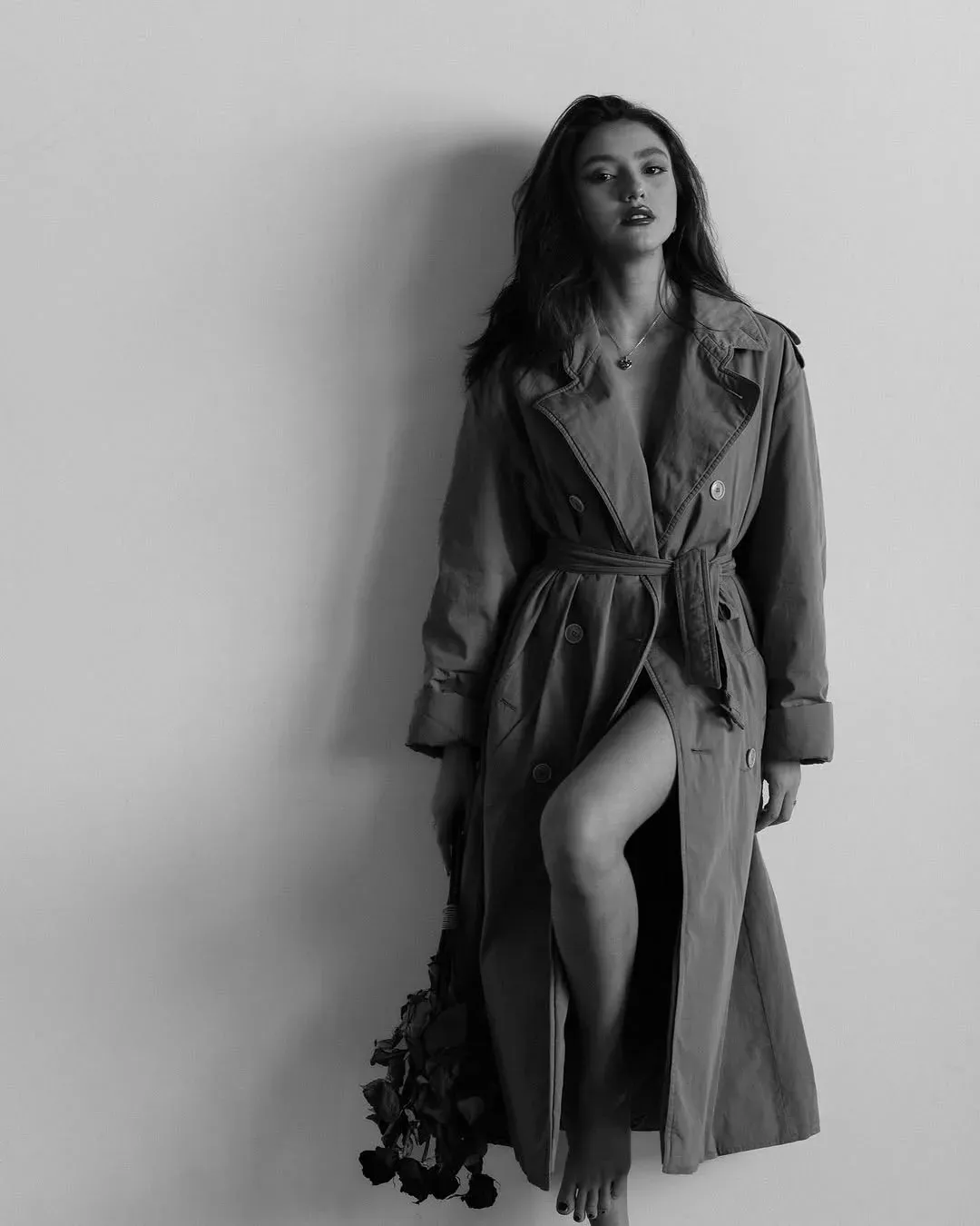 Black and white full-length portrait of a woman in a belted trench coat against a plain wall, holding dried roses at her side.