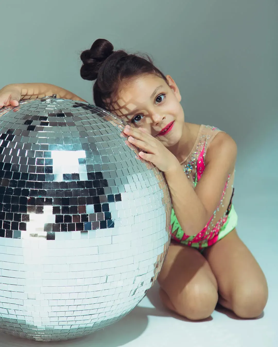 A young gymnast in a sequinned leotard kneels and rests her cheek against a large mirrorball.