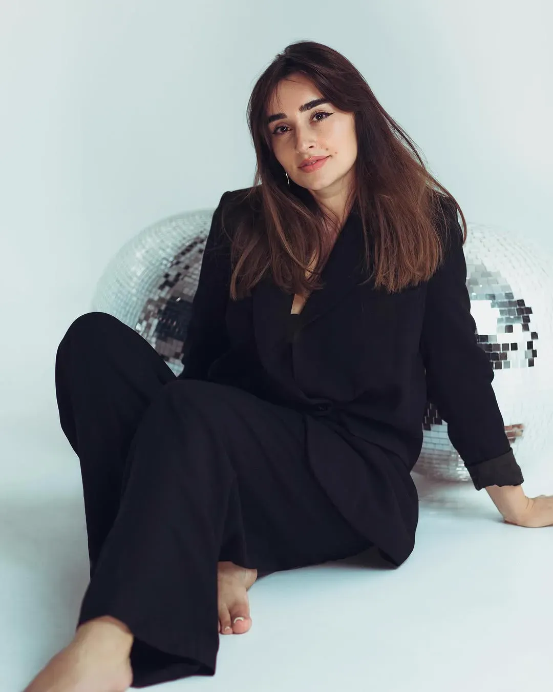 A woman in a loose black suit sits barefoot on a white studio floor between two mirrorballs.
