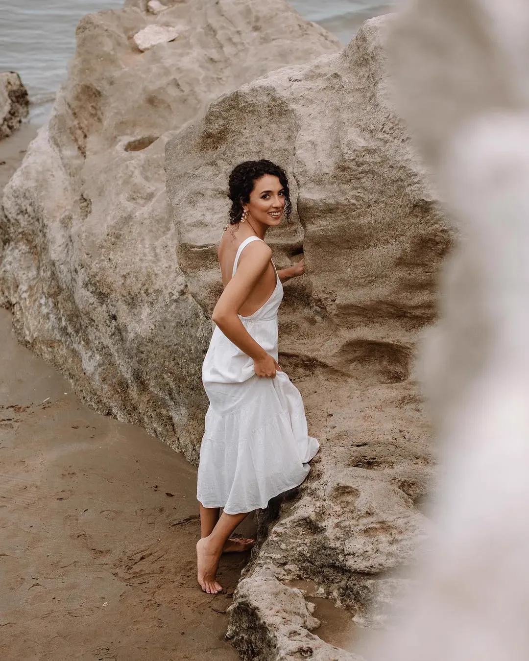 A woman in a white sundress glances back over her shoulder while climbing a pale sandstone rock at the shore.