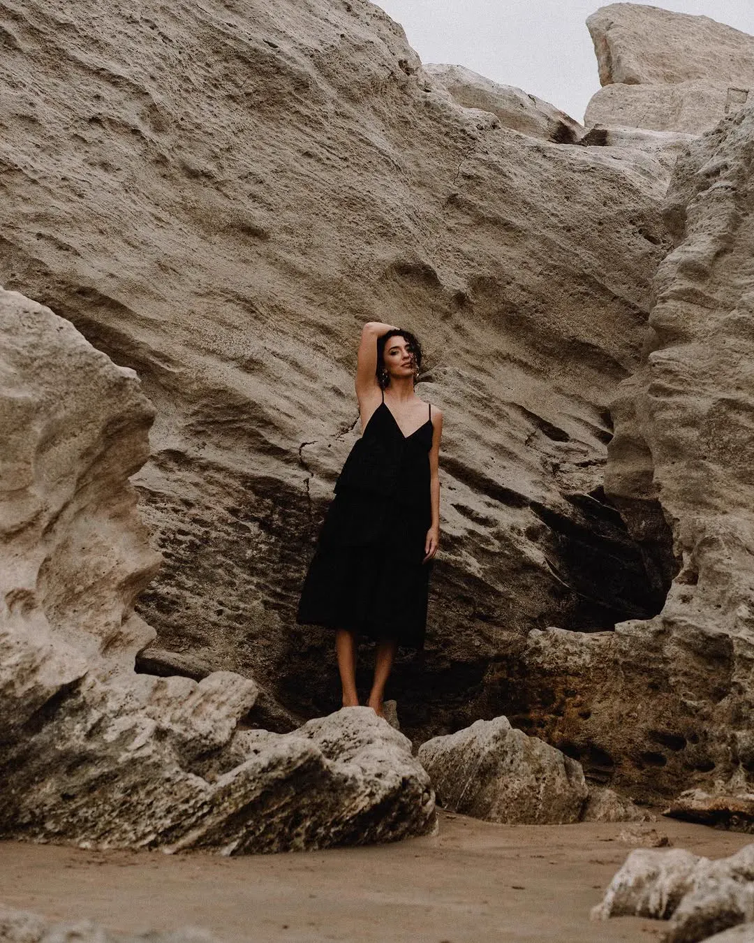 A woman in a black dress stands small against a vast weathered sandstone cliff, one arm raised to her hair.