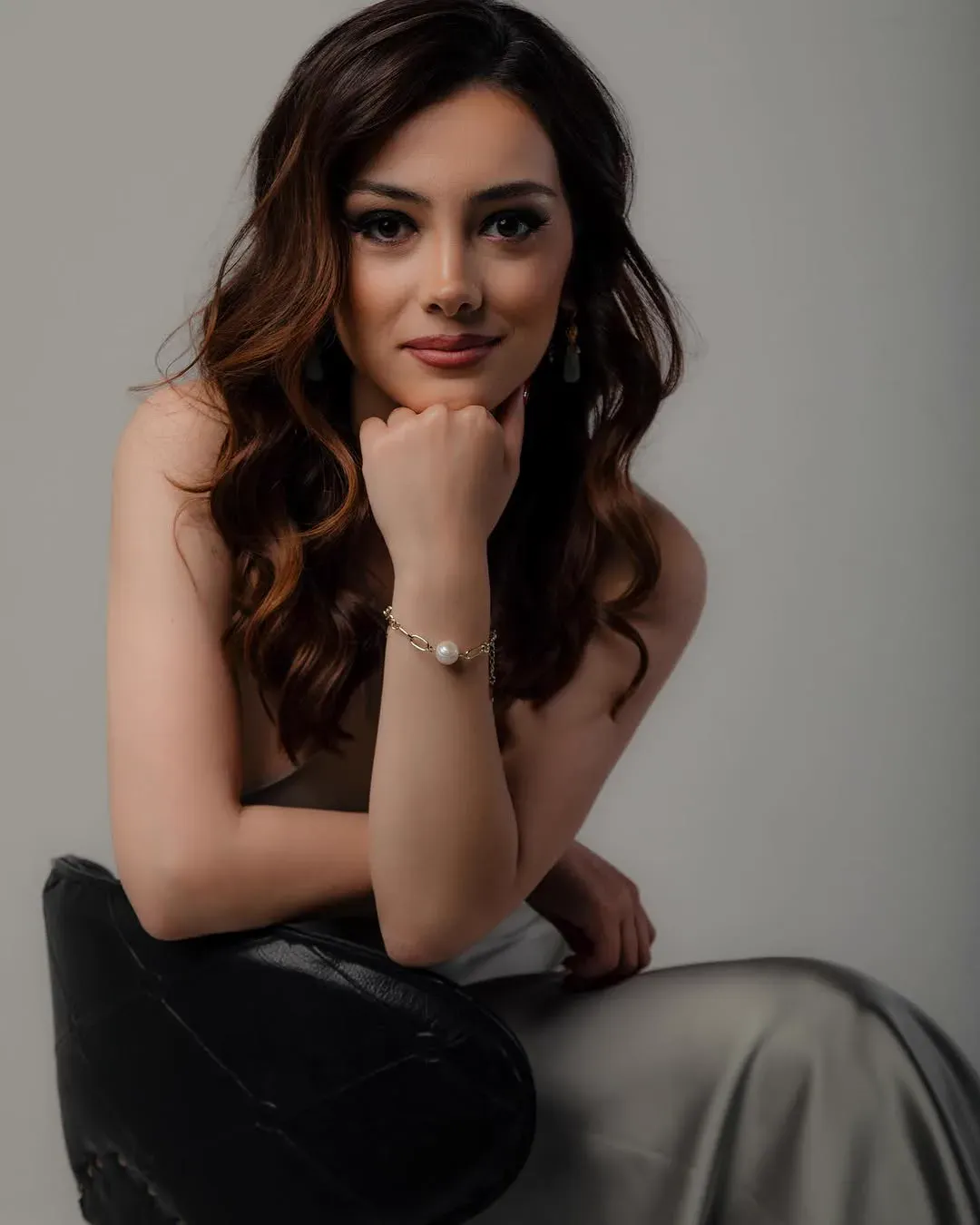 Sada, seated against a soft grey studio backdrop in a satin dress, resting her chin on her fist and looking at the camera.