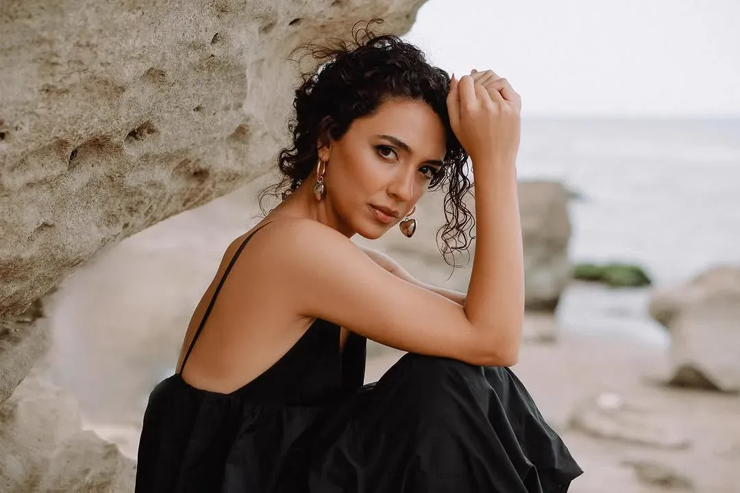 A woman in a black dress sits against sandstone at the waterline, chin resting on her hand, looking at the camera.