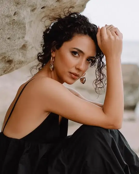 A woman in a black dress sits against sandstone at the waterline, chin resting on her hand, looking at the camera.